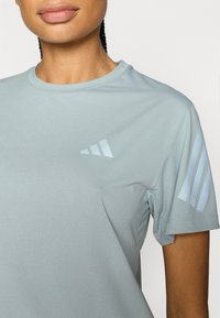 Jasnoniebieska sportowa koszulka wykonana z gładkiej tkaniny. Posiada okrągły dekolt, krótkie rękawy oraz trzy białe pionowe paski na ramieniu.