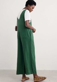 Žalias lininis kombinezonas su plačiomis kojomis, dviem galiniais kišenėmis ir V formos nugaros iškirpte. Derinamas su balta marškinėliais ir rudais šalkais.