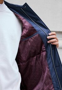 Jachetă de marină cu fermoar, având un interior din satin burgund cu un model embosat. O buzunare de accent albastru este vizibilă în interior.