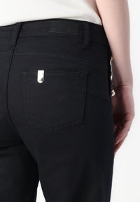 Jean noir avec un tissu texturé, comportant une petite étiquette en argent et une poche arrière de forme triangulaire. Design simple, peu de détails.