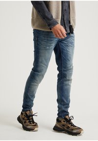 Haalarit siniset slim fit -farkut, joissa on kuluneet yksityiskohdat, yhdistettynä ruskeisiin ja mustiin urheilukenkään, joissa on tekstuurilla varustettuja yksityiskohtia ja nauhoja.
