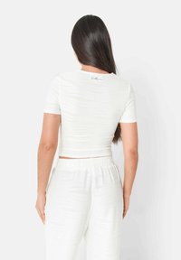 Hvid, tætsiddende cropped top med korte ærmer og et struktureret mønster, der har en rund halsudskæring og et synligt logo-detalje på ryggen.