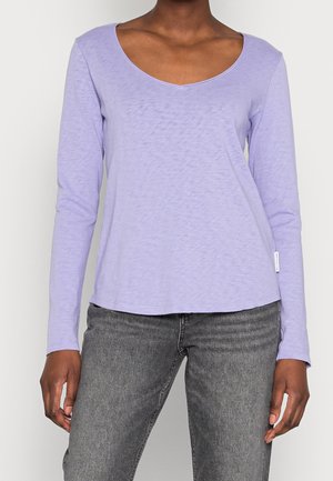 Lila Langarmshirt mit weitem V-Ausschnitt, weicher Textur und geradem Saum. Kombiniert mit dunkelgrauen Jeans; enthält ein kleines gewebtes Etikett am Saum.