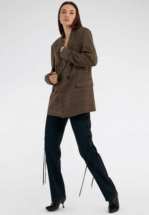 Donna che indossa un blazer oversize a quadri marroni, pantaloni neri con dettagli laterali allacciati e décolleté nere con punta appuntita, in piedi su uno sfondo bianco.