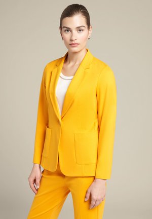 Gelber Blazer mit taillierter Passform, Reverskragen und zwei vorderen Taschen. Über einem weißen Oberteil getragen. Glatte Textur und lebendige Farbe.