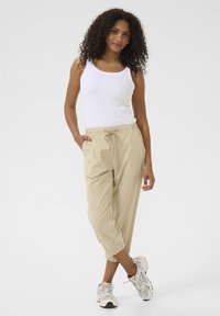Lettere, stribede beige bukser med elastisk talje og snøre, parret med en hvid tanktop og grå sneakers.