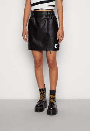 Jupe en cuir noir avec taille froncée, deux poches latérales et un écusson logo brodé. Portée avec des bottines noires à lacets jaunes.