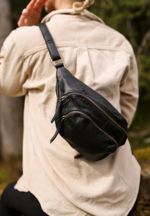 Schwarze Leder-Schultertasche mit zwei Reißverschlussfächern und strukturierter Oberfläche. An der Tasche befestigter Riemen mit einer metallischen Schnalle. Hintergrunde scheint natürlich.