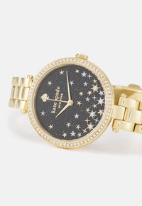 Gold-tone watch with a black starry dial, featuring silver and gold star accents and a crystal-encrusted bezel. Bracelet-style band.