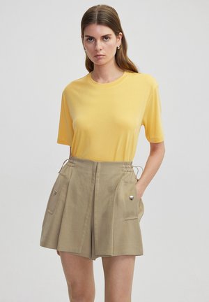 T-shirt en coton jaune associé à un short beige avec taille élastique, poches latérales et accents métalliques brillants. Texture lisse, coupe décontractée.