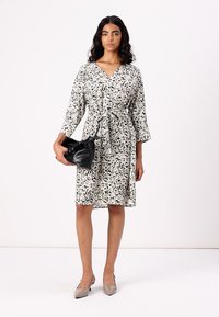White dress with black abstract pattern, tie waist, three-quarter sleeves, worn with a black handbag and taupe high heels.