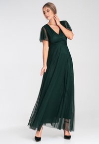 Dunkelgrünes Maxikleid mit V-Ausschnitt, kurzen durchsichtigen Ärmeln und taillierter Taille. Der fließende Stoff erzeugt eine elegante Silhouette. Schwarze High Heels.