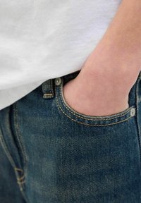 Traperice s opuštenim krojem, s prednjim džepom, kontrastnim žutim šavovima i svijetloplavim pranjem. Obična bijela majica vidljiva iznad.