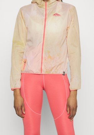 Kobieta ubrana w jasnoróżową i brzoskwiniową kurtkę Nike Trail z suwakiem oraz dopasowane koralowo-różowe legginsy, stojąca na jednolitym tle.