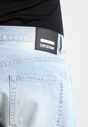 Hellblaue Jeanshose aus Denim mit einem sichtbaren schwarzen rechteckigen Markenlabel mit der Aufschrift "DRDENIM" nahe dem Bund, kombiniert mit einem schwarzen Oberteil.