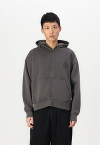 Sweat à capuche gris avec fermeture éclair, poche avant, coupe oversize et cordon de serrage. Il comporte un logo discret et des détails de couture contrastants.
