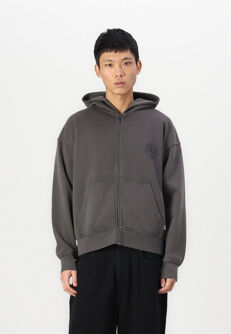 Sweat à capuche gris avec fermeture éclair, poche avant, coupe oversize et cordon de serrage. Il comporte un logo discret et des détails de couture contrastants.
