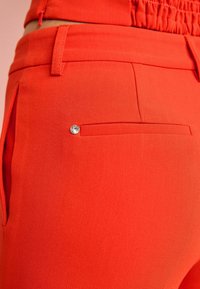 Red tailored trousers featuring smooth fabric, a side pocket with a decorative rhinestone, and an elasticized waistband for comfort.
