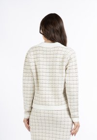 Strick-Cardigan in Creme und Beige mit einem strukturierten Muster. Verfügt über einen gerippten Kragen, Manschetten und Taillenbund. Wird über einem passenden Rock getragen.