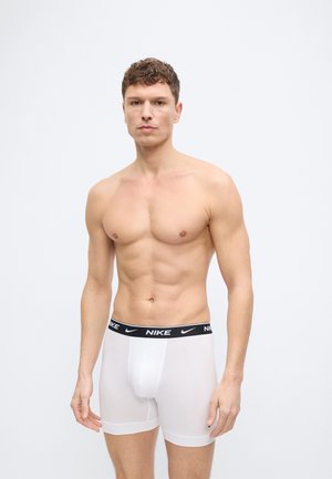 Ustrezen mlad moški s kratkimi kodrastimi lasmi, oblečen v bele Nike boksarice s črnim pasom z napisom blagovne znamke, stoji pred svetlo enobarvno steno.