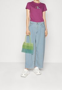 Fialové tričko s krátkým rukávem a logem, světle modré široké džíny, zelená taška na korálky s gradientem a bílé dřeváky se větracími dírami.