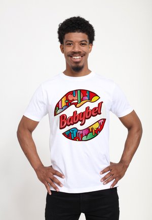 White T-shirt featuring a circular, colorful Babybel logo with drippy patterns. Model wears black pants, hands on hips, smiling.