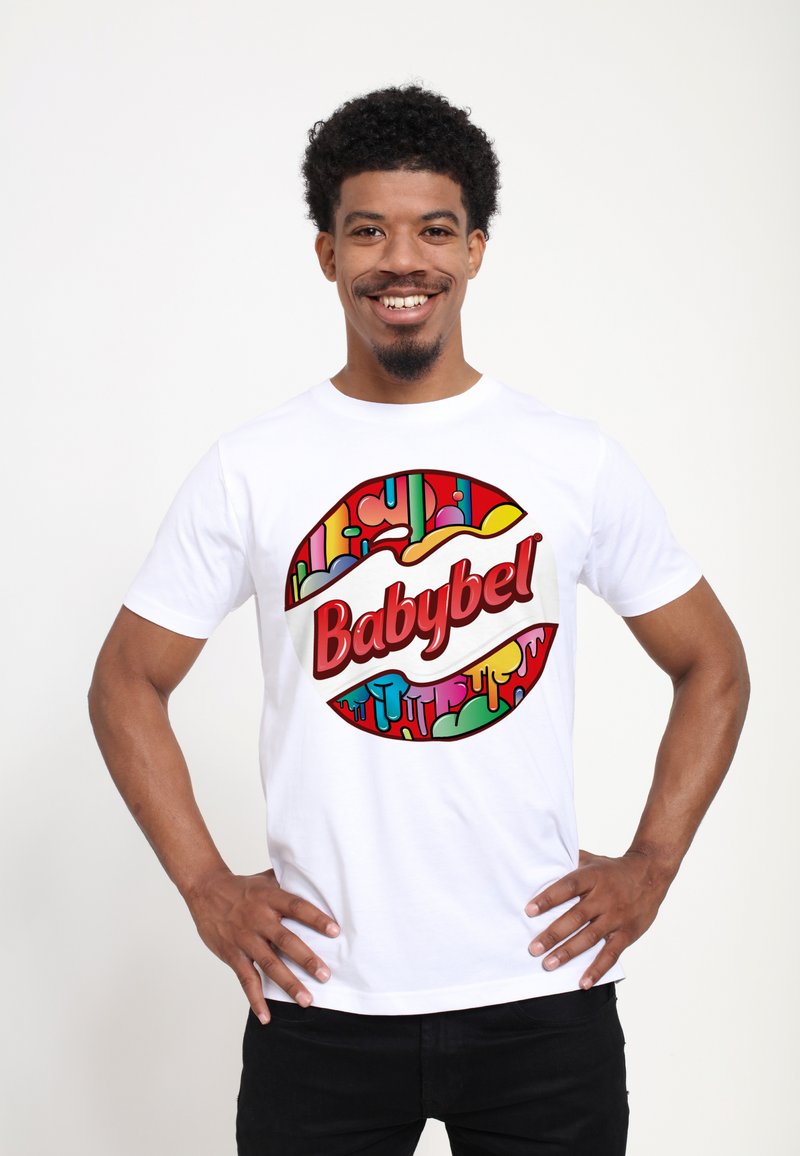Bílý tričko se zaobleným, barevným logem Babybel a kapajícími vzory. Modelka má na sobě černé kalhoty, má ruce na bocích a usmívá se.