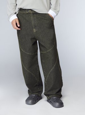 Pantaloni in denim verde con cuciture gialle, vestibilità ampia e design con pannelli curvi. Abbinati a zoccoli neri; il modello indossa una maglietta grigia a maniche lunghe.