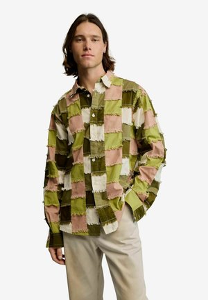Junger Mann mit schulterlangem Haar, der ein langärmeliges Patchwork-Hemd in Grün-, Braun-, Weiß- und Rosatönen trägt, kombiniert mit beigen Hosen.