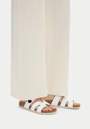 Białe sandały z wycięciami, płaską korkową podeszwą i regulowanym paskiem. Noszone z jasnym, zwiewnym materiałem.