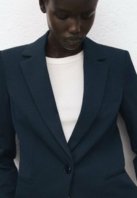 Marineblauw colbert met een getailleerd ontwerp, sluiting met één knoop en klepzakken, gedragen over een wit geribd shirt. Soepele stoftextuur.