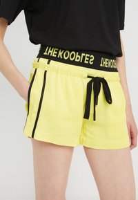 Pantaloni corti gialli con accenti neri, cintura elasticizzata e cordoncino. Presentano tasche laterali e una texture liscia e leggera.
