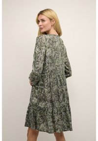 Femeie blondă îmbrăcată într-o rochie lungă cu mâneci, cu model paisley verde, întoarsă ușor cu mâinile aproape de talie, pe un fundal simplu.
