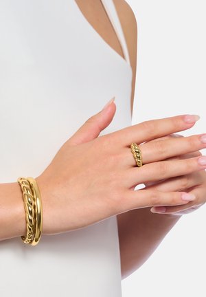 Weibliche Hände mit natürlichen Nägeln, die einen goldenen Kettenglied-Ring und zwei passende goldene Armbänder über einem weißen ärmellosen Oberteil tragen.