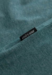 Tyrkysová látka s texturovaným povrchem, která má malou černou nálepku s bílým textem "ombre wear."