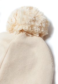 Beige Strickmütze mit einem flauschigen Bommel oben. Glatte Textur, runde Form und ein einfaches, stimmiges Design ohne sichtbare Muster.