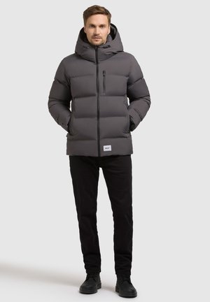 Graue gepolsterte Jacke mit Kapuze, Reißverschluss und vorderer Reißverschlusstasche. Getragen über schwarzen Hosen und Schuhen. Glatte Textur, minimales Design.