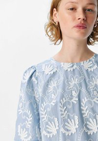 Blusa azul clara con bordado floral blanco, mangas abullonadas y escote redondeado, hecha de una tela suave con un detallado intrincado.