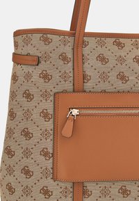 Sac fourre-tout beige en tissu texturé avec un motif répétitif, agrémenté d'accents en cuir marron et d'une poche zippée.