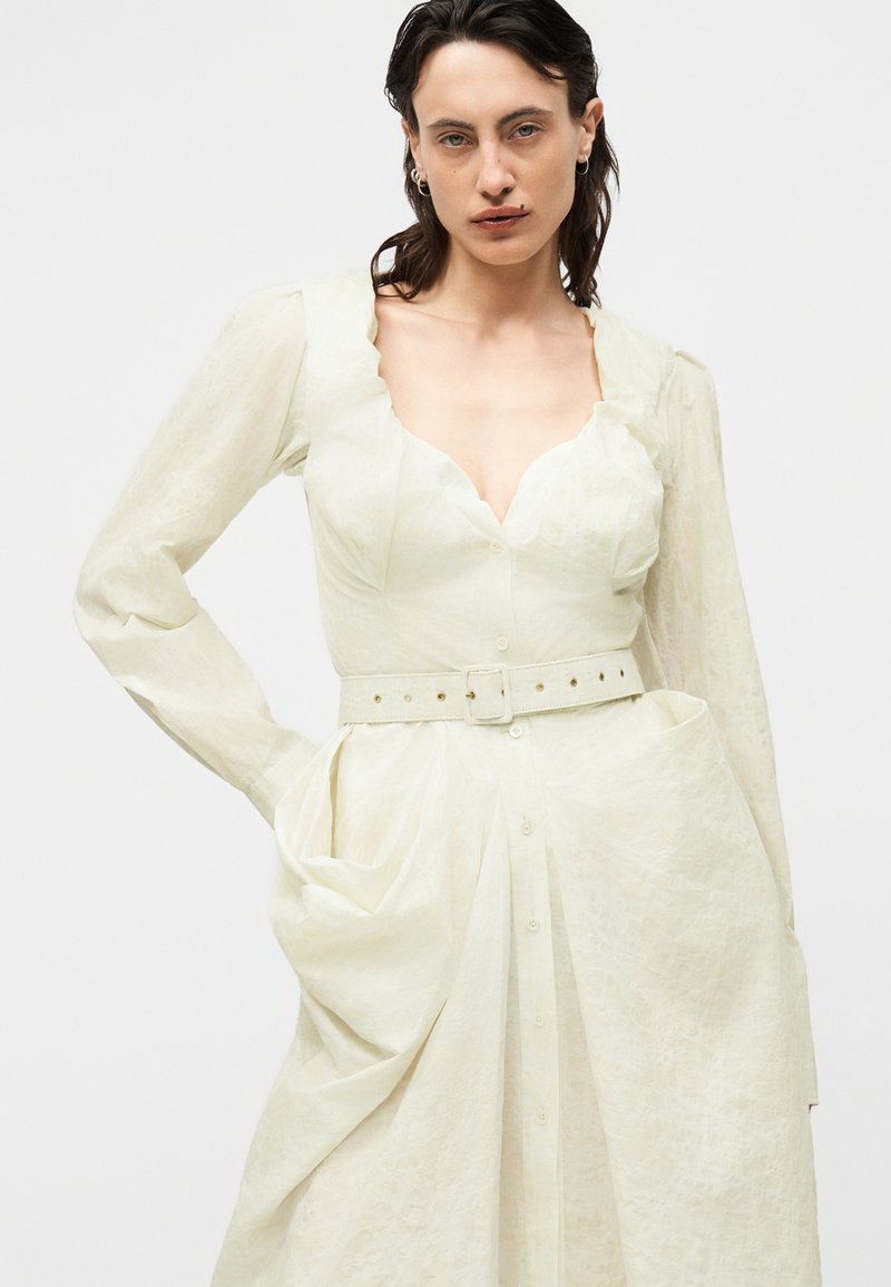 Femme portant une robe crème ceinturée, avec un col festonné et des manches longues, posant avec une main dans la poche devant un fond uni.