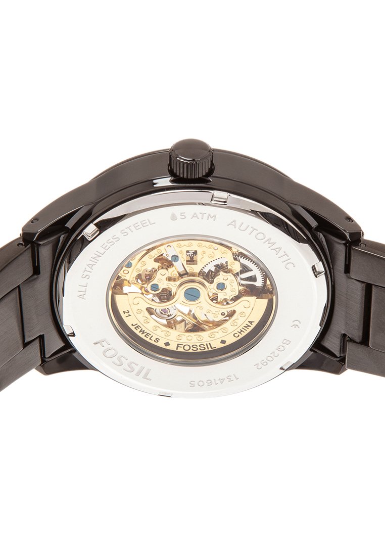 Parte posterior de un reloj automático Fossil de acero inoxidable negro que muestra engranajes expuestos de tono dorado y el texto "27 joyas", "5 ATM" y "China".