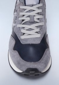 Grauer sportlicher Sneaker aus einer Kombination von Wildleder und Leder, mit Schnürverschluss und einer weißen Mittelsohle sowie einer strukturierten dunklen Sohle.