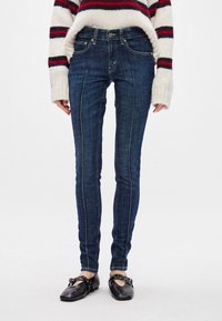 Ajuda azul escuro slim com costura visível, design de cinco bolsos e cintura média, combinada com sapatos pretos planos.