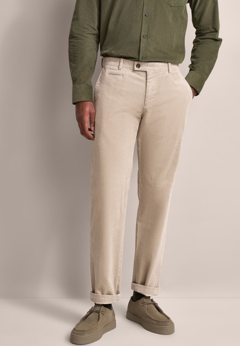 Beige Cord-Hosen mit geradem Schnitt, Fronttaschen und aufgerollten Säumen, kombiniert mit einem grünen Hemd mit Knöpfen und grauen Schuhen.