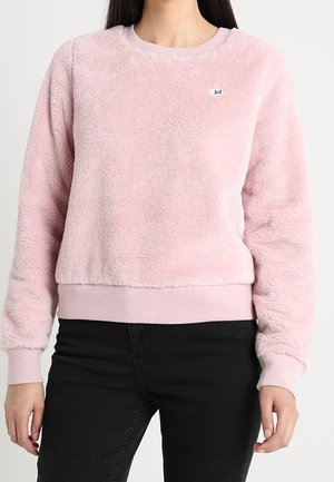 Osoba ubrana w jasnoróżowy, miękki sweter z długim rękawem, z prążkowanymi mankietami i pasem, w połączeniu z czarnymi spodniami.