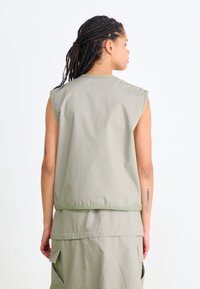 Bezramenná vesta v svetle olivové barvě z voděodolné tkaniny, s rovnými okraji, minimálním šitím a kontrastn�ím zeleným lemováním.