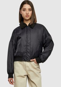 Jachetă bomber neagră, scurtă, cu guler pufoasă, design căptușit, manșete elastice și buzunare laterale, combinată cu pantaloni bej deschis.