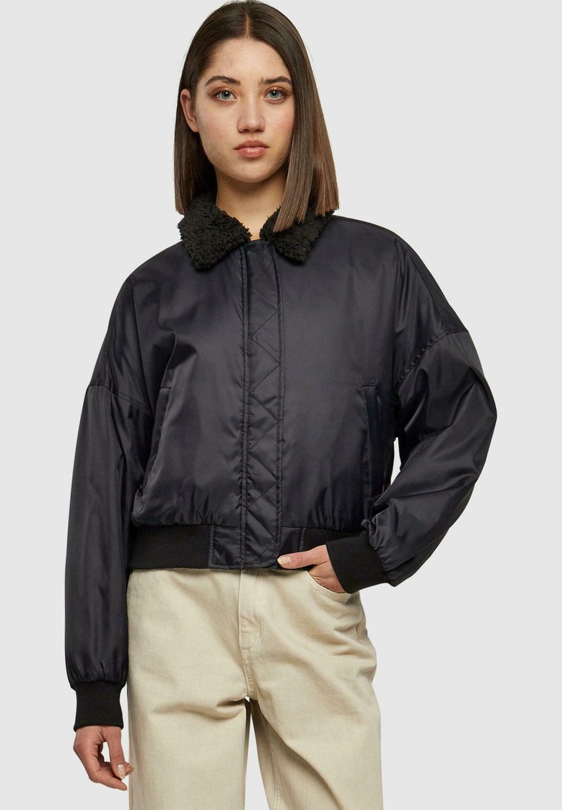 Jachetă bomber neagră, scurtă, cu guler pufoasă, design căptușit, manșete elastice și buzunare laterale, combinată cu pantaloni bej deschis.