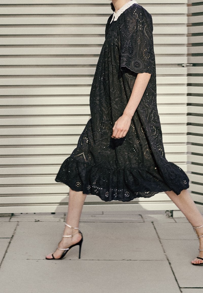 Czarna koronkowa sukienka z krótkimi rękawami, ozdobiona szczegółowym haftem i falbanistym dołem, zestawiona z sandałkami na wysokim obcasie z paskami.