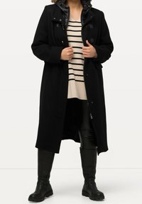 Black long coat with a quilted collar, featuring toggle closures. The model wears a striped sweater and black shiny leggings with knee-high boots.